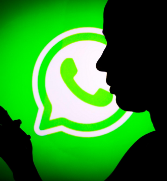 Persona a contraluz escribe en celular. A fondo se ve logo de WhatsApp